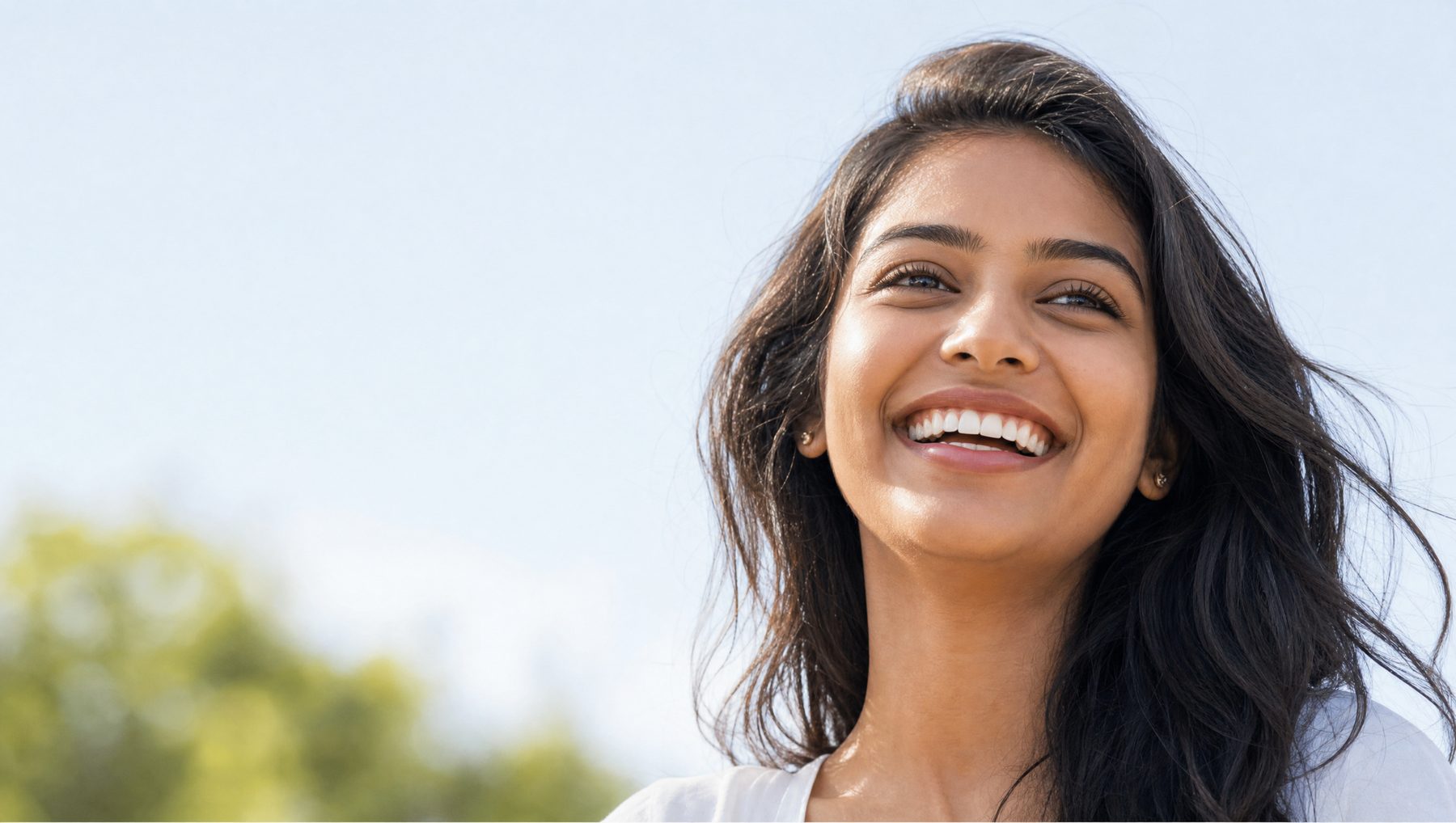 A young woman laughing, looking up, against an open pale-blue sky.